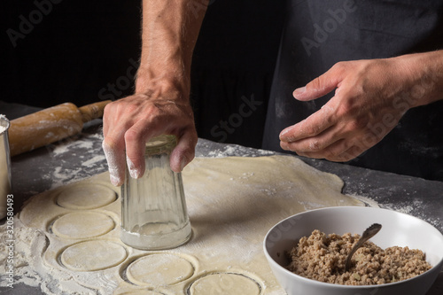 A man is making a dough for varenyky or dumplings, with meat, onions and bacon. Man in a black apron without face. Man squeezes out circles of dough with a glass. Making dough by male hands at bakery 