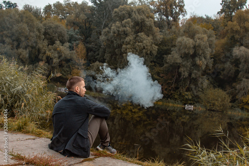 A guy with a beard sits on the lake and smokes an electronic cigarette. Blows puffs of smoke. The concept of unity with nature. Rest near the water. Vape, Man Vaping. lips and smoke, a lot of smoke. © Yuliia