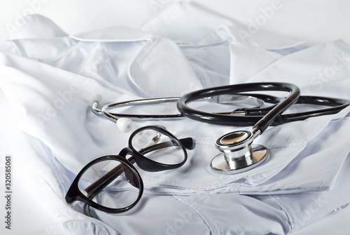 Black stethoscope on white medical gown. Black round glasses near medical equipment.