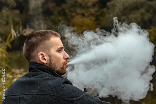 A guy with a beard sits on the lake and smokes an electronic cigarette. Blows puffs of smoke. The concept of unity with nature. Rest near the water. Vape, Man Vaping. lips and smoke, a lot of smoke.