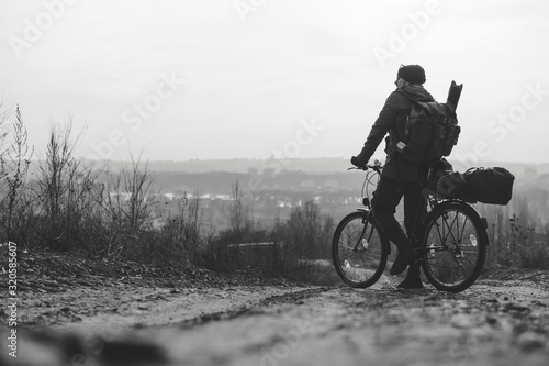 A rider man with a backpack and a bicycle stands and looks into the distance. hilltop overlooking a valley in haze, a city on the horizon. winter or autumn landscape road. Black and white