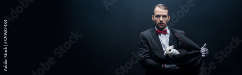 panoramic shot of professional magician holding white rabbit in hat, dark room with smoke