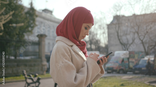 Pretty young Muslim lady using a smartphone on the lovely street in modern city. Busy female traveler in hijab using mobile phone.