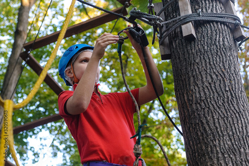 A teenage boy climbs in a rope park. There is a protective helmet on his head. The boy has special climbing equipment. Summer.
