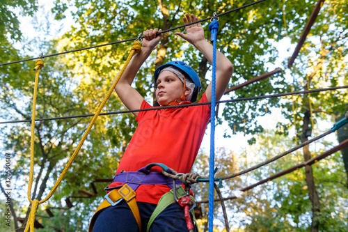 A teenage boy climbs in a rope park. There is a protective helmet on his head. The boy has special climbing equipment. Summer.
