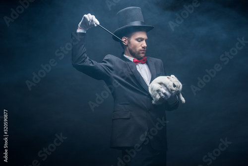 emotional magician in suit and hat showing trick with wand and white rabbit, dark room with smoke