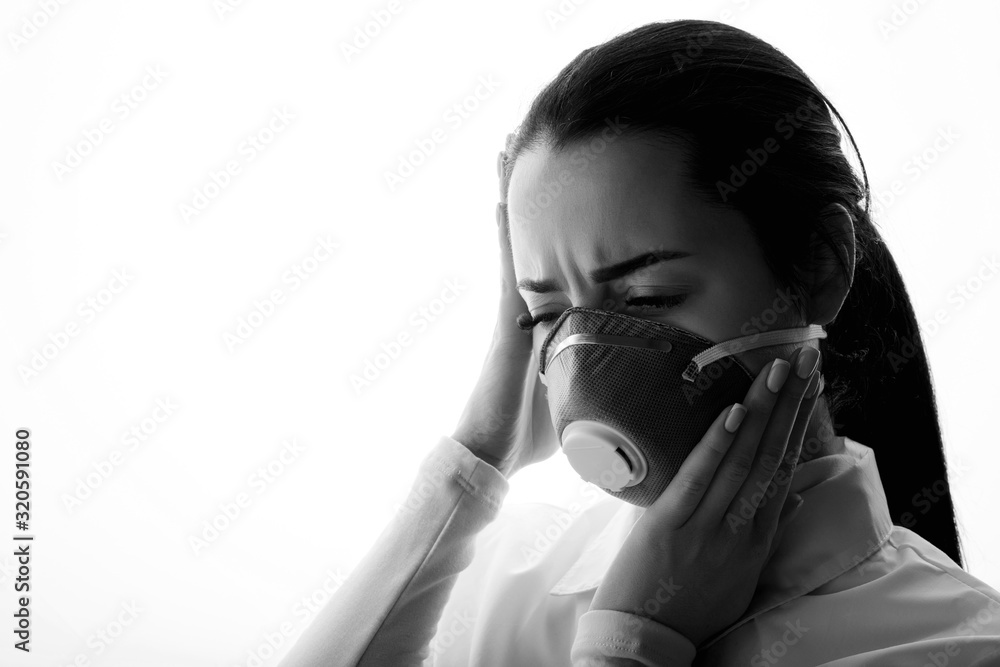 Black and white portrait of young sad female doctor suffering from severe headache, pressing her fingers to temples. Woman in protective respirator. Free space for text. Healthcare concept. Black and white portrait of young sad female doctor suffering from severe headache, pressing her fingers to temples. Woman in protective respirator. Free space for text. Healthcare concept.