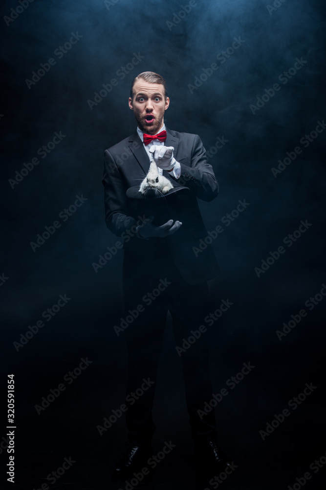 excited magician in suit showing trick with white rabbit in hat, dark room with smoke excited magician in suit showing trick with white rabbit in hat, dark room with smoke