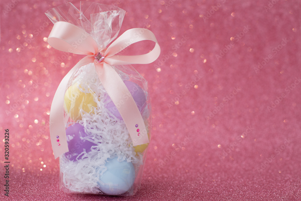 Multi-colored purple, pink, blue, yellow Easter eggs in a transparent gift package, decorated with a bow and rhinestones, on a pink shiny bokeh background. Symbol of celebration of religious holiday Multi-colored purple, pink, blue, yellow Easter eggs in a transparent gift package, decorated with a bow and rhinestones, on a pink shiny bokeh background. Symbol of celebration of religious holiday