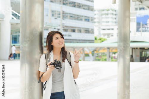Charming Asian woman in gray shirt and open shirt carrying a backpack and holding camera, make a finger point on the air from right side with urban office building background. Free Download Charming Asian woman in gray shirt and open shirt carrying a backpack and holding camera, make a finger point on the air from right side with urban office building background. - GFXLoad