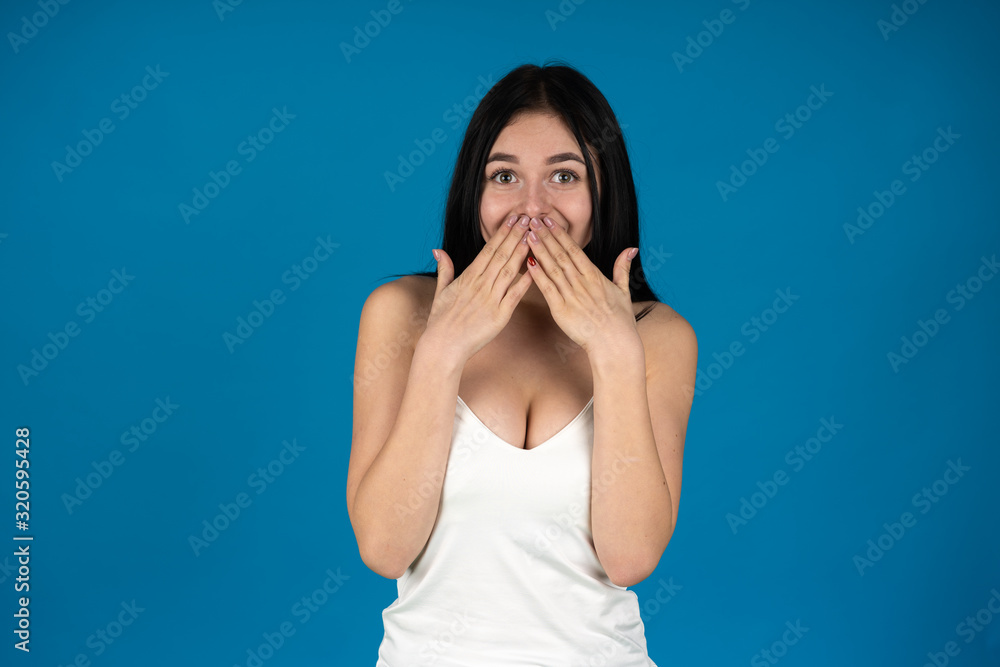 Front view of shocked brunette girl covering mouth with hands isolated on blue background Front view of shocked brunette girl covering mouth with hands isolated on blue background