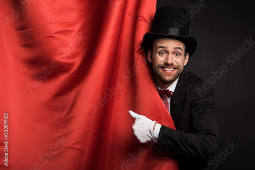 smiling magician in suit and hat showing thumb up in circus with red curtains