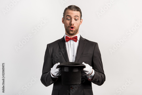 surprised magician in gloves looking into hat, isolated on grey photo