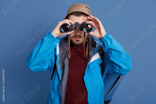 Horizontal studio picture of experienced hiker looking through binoculars, go camping alone, standing isolated over phantom blue background, wearing blue jacket, hat and dark red sweatshirt. photo