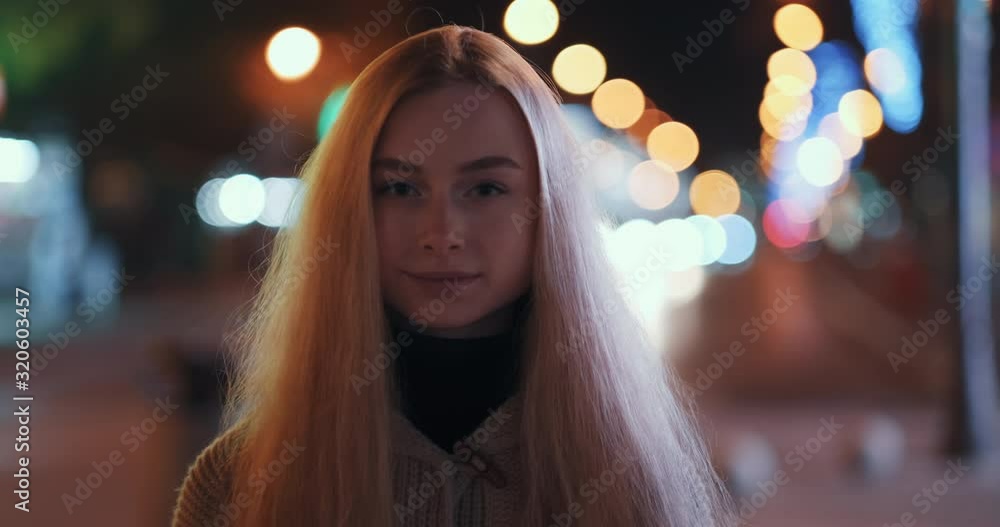 Portrait of happy blonde girl with long hair stands and smiles, looks at camera against of passing cars and bright bokeh of city lights in evening in winter after walk. Emotions of people. Lifestyle Portrait of happy blonde girl with long hair stands and smiles, looks at camera against of passing cars and bright bokeh of city lights in evening in winter after walk. Emotions of people. Lifestyle