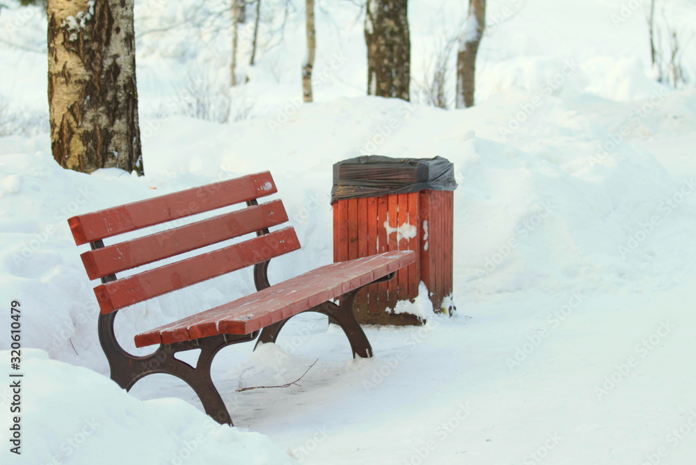 Wooden red bench on black metal base in park on background of snowdrifts and trees.