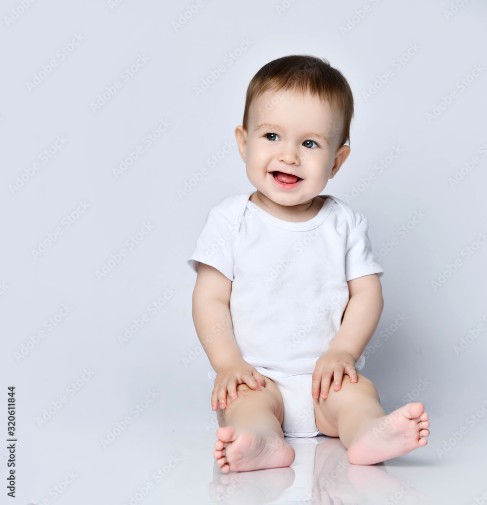Happy smiling laughing baby boy toddler in white bodysuit is sitting on the floor looking sideways at copy space on gray Happy smiling laughing baby boy toddler in white bodysuit is sitting on the floor looking sideways at copy space on gray
