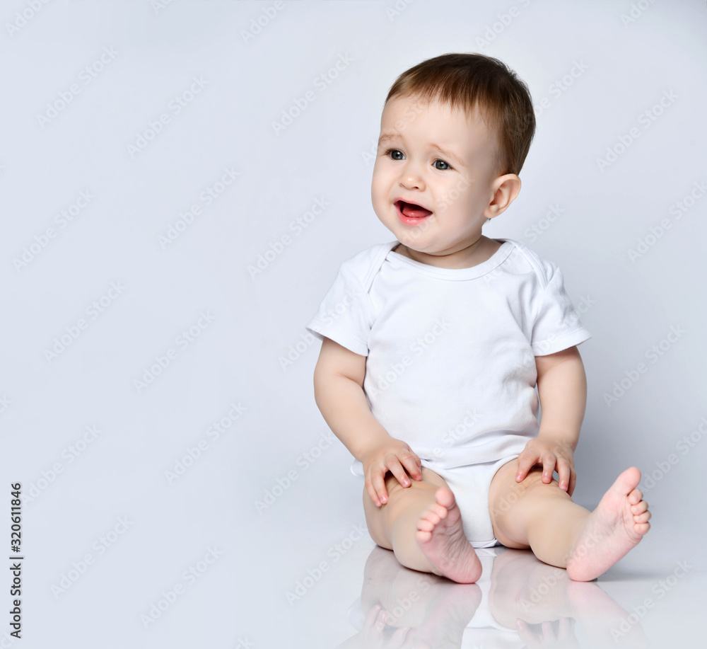 Upset barefooted baby boy toddler in white bodysuit is sitting on the floor looking sideways going to cry Upset barefooted baby boy toddler in white bodysuit is sitting on the floor looking sideways going to cry