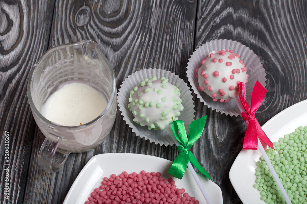 Cake pops in white glazed chocolate with green and pink sprinkles. A green and pink bow is tied on sticks. Nearby melted chocolate and topping in plates. Against the background of brushed pine boards Cake pops in white glazed chocolate with green and pink sprinkles. A green and pink bow is tied on sticks. Nearby melted chocolate and topping in plates. Against the background of brushed pine boards