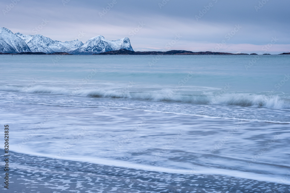 Arctic landscape with long exposure Waves in foreground rolling to coast and snowy mountains in background on a cloudy winter day, Skaland, Senja, Norway