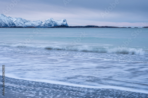 Arctic landscape with long exposure Waves in foreground rolling to coast and snowy mountains in background on a cloudy winter day, Skaland, Senja, Norway