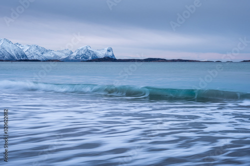 Arctic landscape with long exposure Waves in foreground rolling to coast and snowy mountains in background on a cloudy winter day, Skaland, Senja, Norway © sg-naturephoto.com 