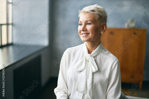 Photographie Attractive middle aged lady in stylish white silky blouse thinking about something pleasant, sitting by window in office interior, rejoicing at good day, ready to do work routine
