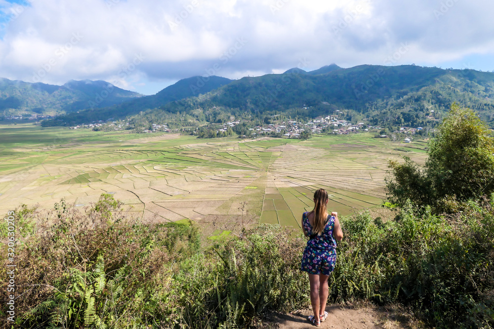 A woman in a jumpsuit admiring the colorful rice paddies forming a giant spider web in Ruteng, on island of Flores, Indonesia. There are mountains around the paddies. Lingko Spider Web Rice Fields