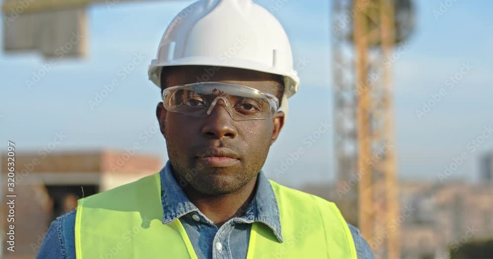 Close up of the African American young handsome man builder in hardhat and goggles looking at the camera and smiling. Outdoor at the building site. Close up of the African American young handsome man builder in hardhat and goggles looking at the camera and smiling. Outdoor at the building site.