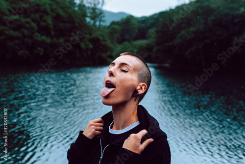 Short haired female with piercing wearing black hoodie looking up while catching raindrops with tongue near green forest and pond