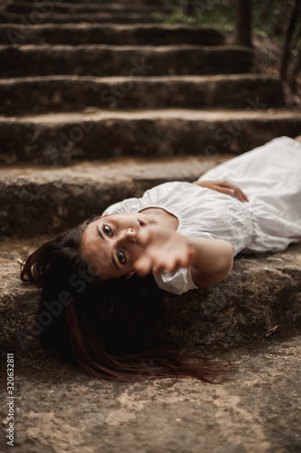 From above of tender charming young female in white dress lying down vulnerably on stairs in autumnal park looking at camera reaching with hand