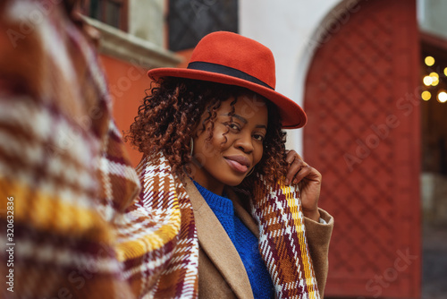 Outdoor fashion portrait of young happy smiling african american woman with afro hairstyle wearing stylish orange hat, checkered scarf, posing in street of city. Copy, empty space for text