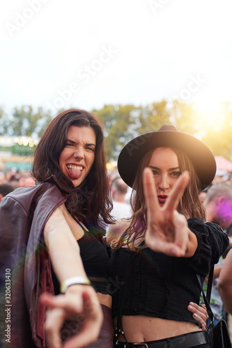 Stylish long haired women in hat and leather jacket grimacing sticking out tongue and showing two fingers at camera in bright day at festival