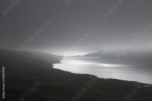 Horizontal low key image with view in the evening light to lake Sørvágsvatn or Leitisvatn lake on Island Vágar of the Faroe Islands covered by fog and North Atlantic Ocean in the background.