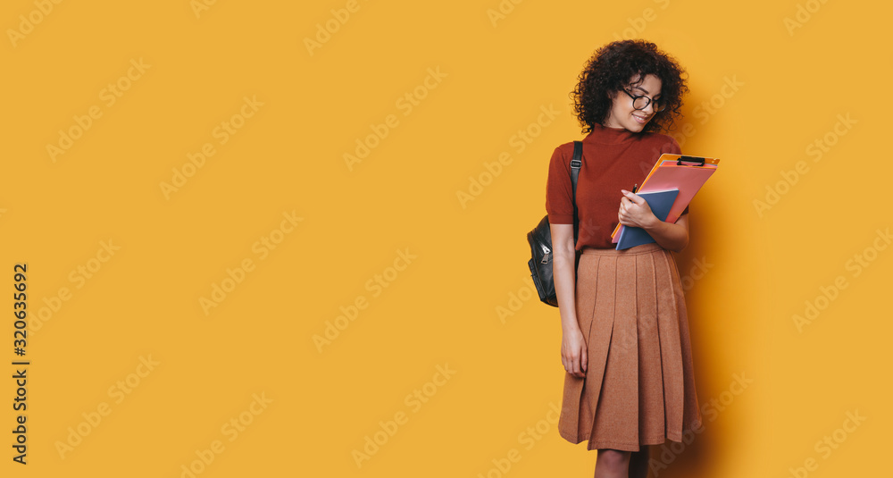 Charming young curly female student looking at her studying books isolated on yellow background.