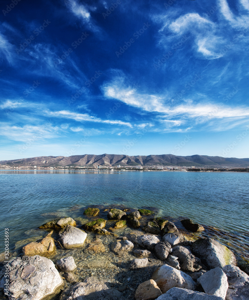 Autumn panorama of Gelendzhik Bay. Clear air, clear sky, and sharp clarity. In the foreground are rocks and a beach. In the background the Caucasus mountains