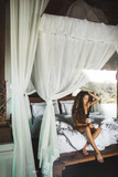 Slim tan woman waking up and enjoying morning coffee in traditional wooden asian bed with white canopy. Wearing black lingerie. Good lazy morning in vacations. 