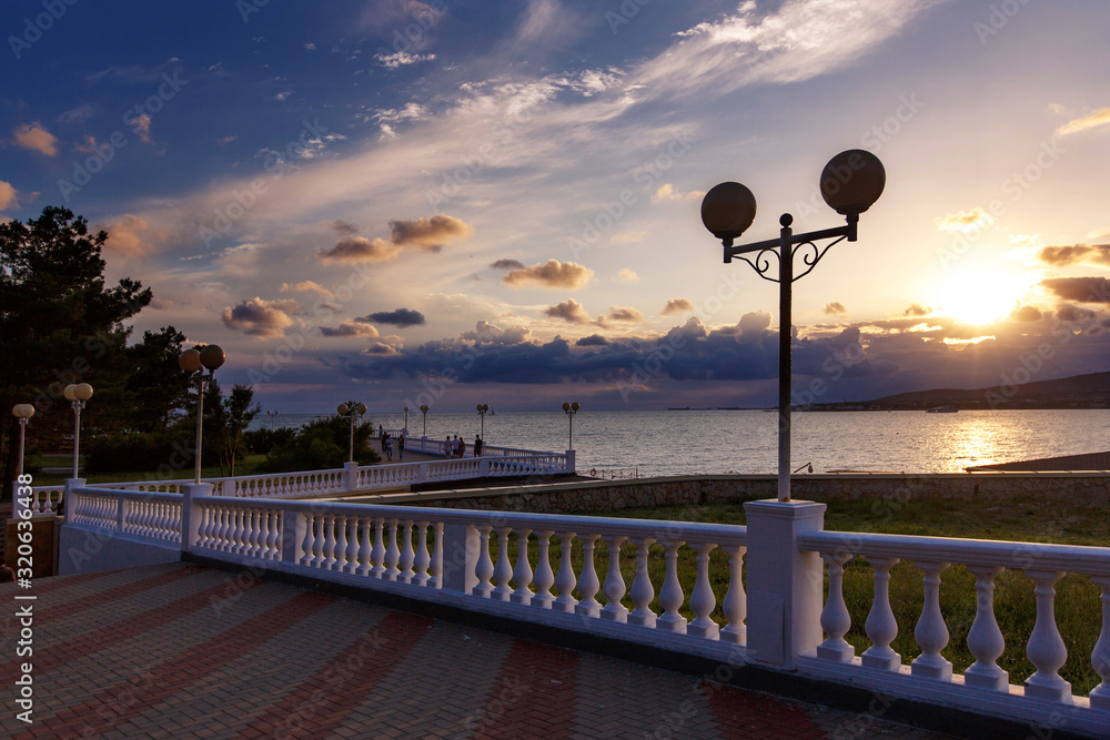 Gelendzhik embankment at sunset. In the foreground is a balustrade that extends into the distance, and lampposts. The sun sets in the sea in the clouds