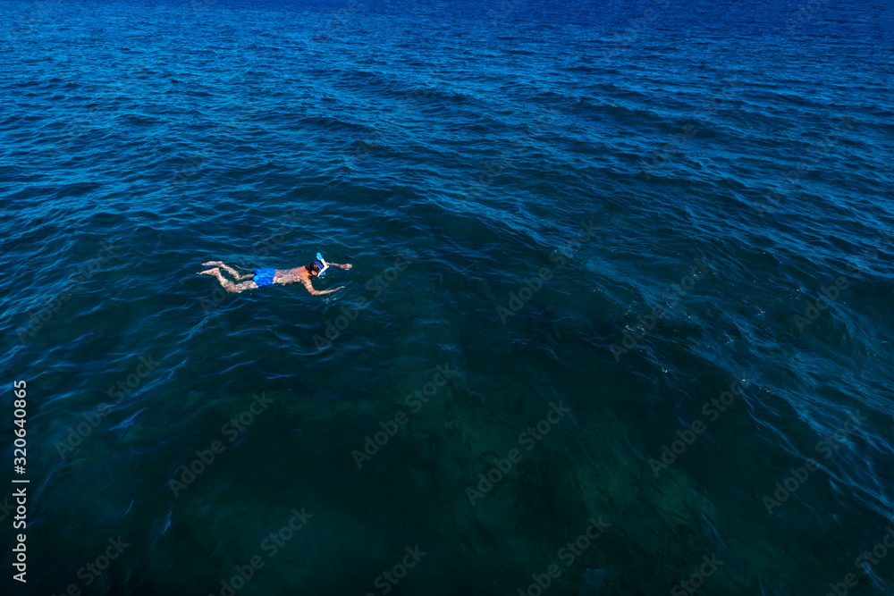 Young man in swimwear lying on blue water. Man in swimming mask relaxing in sea. Tropical summer vacation. Tranquility and peace. Person floating in ocean and watching fish. Stock photo.