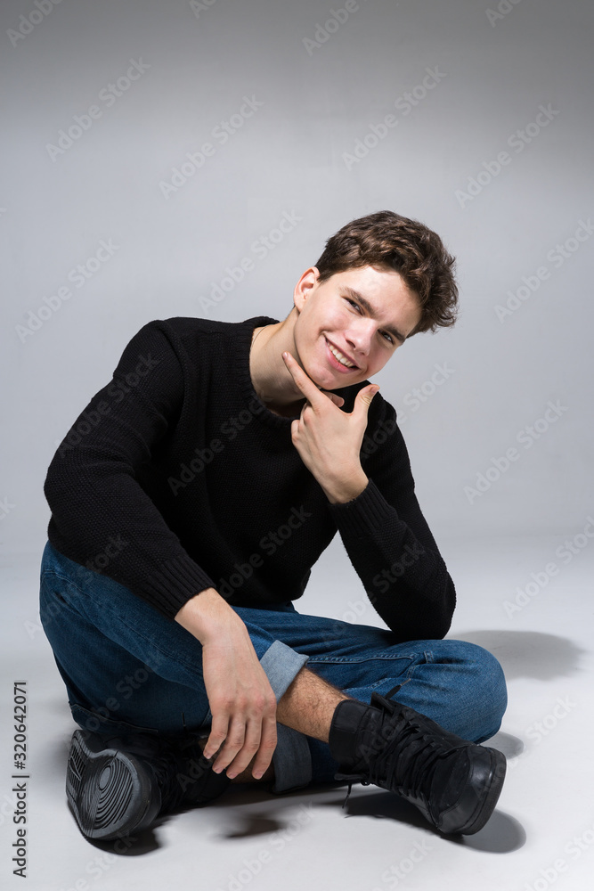 Stylish guy posing in studio sitting on the floor in denim pants and black sweatshirt. Male student portrait full on white background. Studio shot Handsome attractive european man sitting on floor Stylish guy posing in studio sitting on the floor in denim pants and black sweatshirt. Male student portrait full on white background. Studio shot Handsome attractive european man sitting on floor