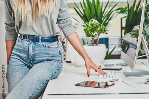 Woman in jeans is sitting on the white table in the office with green plants.