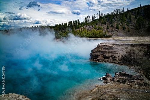 Steam from blue water geyser surrounded by rocks in Yellowstone National Park with puffy clouds and pine trees