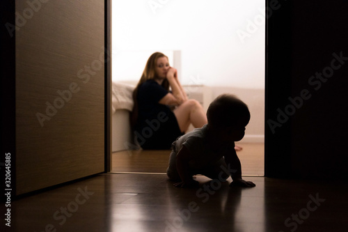 The concept of maternal burnout, postpartum depression, regret and unjustified expectations. Tired young mother sits in the bedroom on the floor, a half-year-old baby crawls nearby. photo