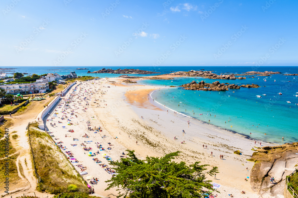 Aerial view over the white sandy beach of Greve Blanche in Tregastel, northern Brittany, France, on a sunny summer day with dozens of people sunbathing and enjoying the turquoise sea.