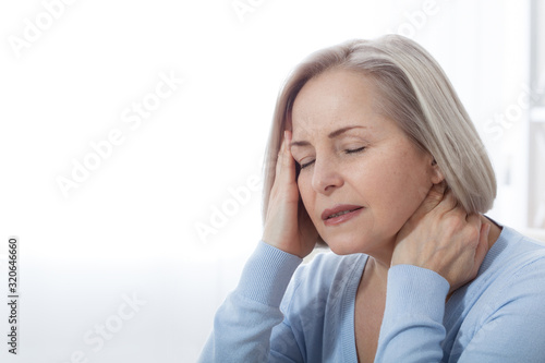 Woman suffering from stress or a headache grimacing in pain as she holds the back of her neck with her other hand to her temple, with copyspace. Concept photo with indicating location of pain. © missty