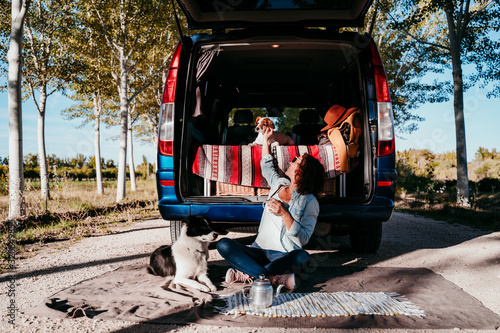 young beautiful woman drinking coffee or tea camping outdoors with a van and her two dogs. Travel concept