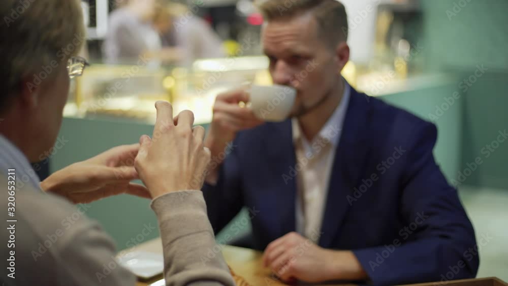 Over the shoulder view tracking shot of middle aged businessman explaining points to business partner drinking coffee during meeting at cafe table Over the shoulder view tracking shot of middle aged businessman explaining points to business partner drinking coffee during meeting at cafe table