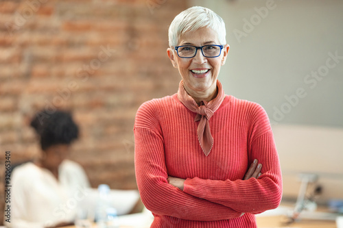 Close-up portrait of mature business woman standing at office. Pretty older business woman, successful confidence with arms crossed in financial building. Mature female in office with team photo