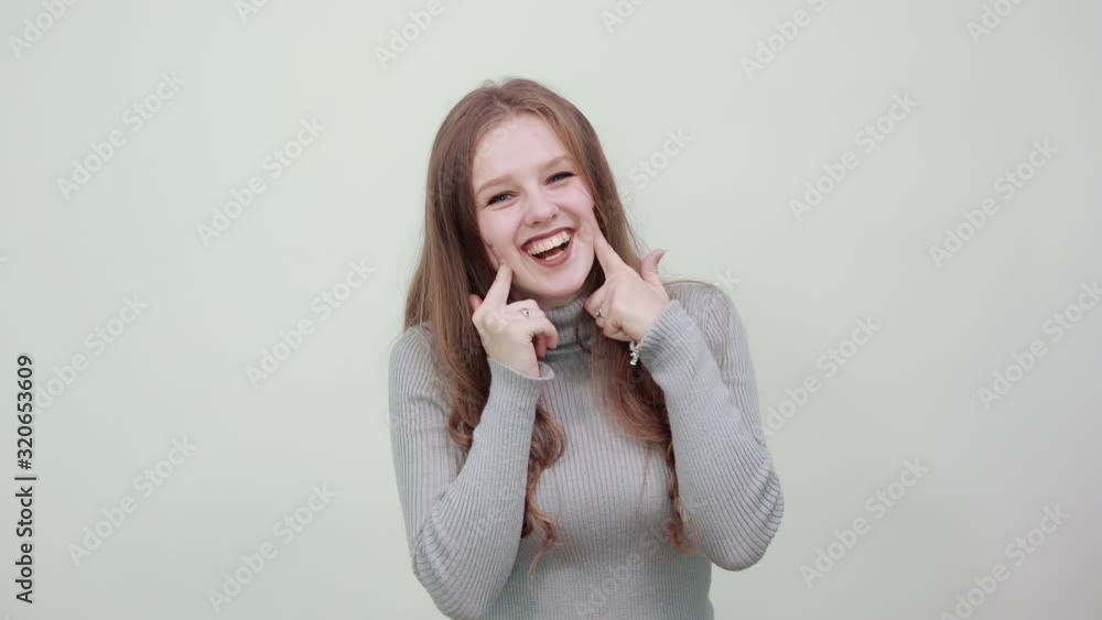 a young beautiful red-haired woman in gray sweater is happy and makes holes in cheeks with index fingers, smiling a young beautiful red-haired woman in gray sweater is happy and makes holes in cheeks with index fingers, smiling