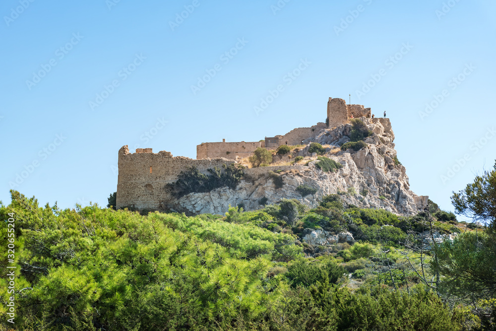 Ruins of medieval castle Kritinia on hill, green branches of pine trees (Rhodes, Greece)